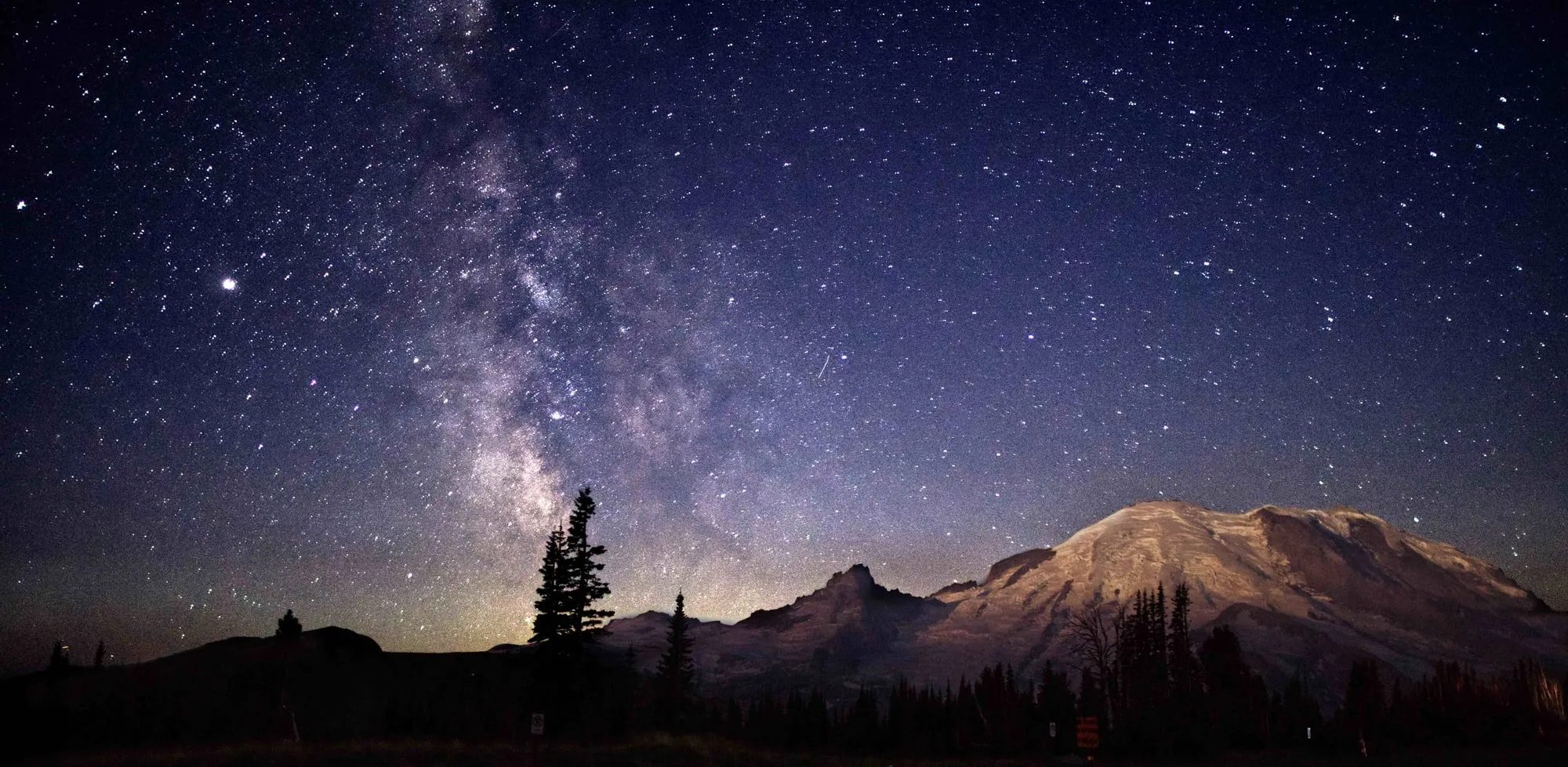 The Milky Way arching beside the silhouette of Mt. Rainier under a star-filled sky.