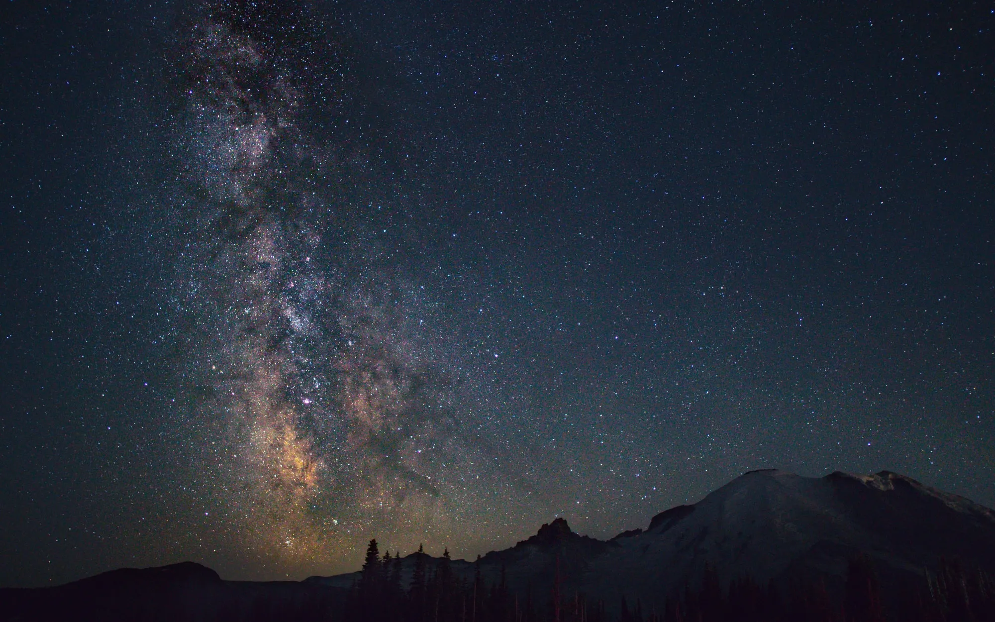 A stacked Milky Way photo rising above Mt. Rainier with pine trees in the foreground.